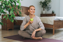 Beautiful Blonde Young Woman In Comfortable Sport Wear Practice Yoga Hold Green Apple In Hand In Cozy Bedroom At Home