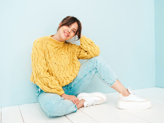 Wall Mural - Young beautiful smiling female in trendy summer yellow hipster sweater and  jeans. Sexy carefree woman posing near light blue wall in studio. Positive model having fun indoors