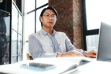 Wall Mural - Vietnamese manager with glasses sits at the table and works at the laptop against the background of large windows