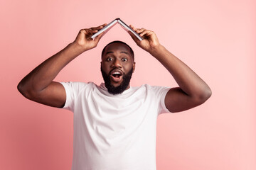 Wall Mural - Portrait of african american man hold notepad roof form above head isolated on pink background