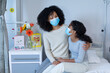 © wavebreak3 - Mixed race mother embracing ill daughter sitting on hospital bed, both wearing face masks