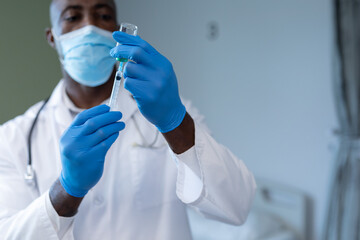 Wall Mural - African american male doctor wearing face mask and gloves preparing covid vaccination