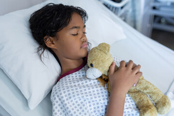 Wall Mural - Mixed race sick girl holding teddy bear lying asleep in bed in a hospital ward