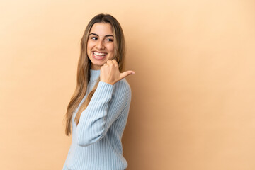 Wall Mural - Young caucasian woman isolated on beige background pointing to the side to present a product