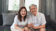 © tirachard - Asian elderly couple using smartphone video conference with grandchild while lying on sofa in living room at home. Enjoying time lifestyle senior family at home concept. Portrait looking at camera.