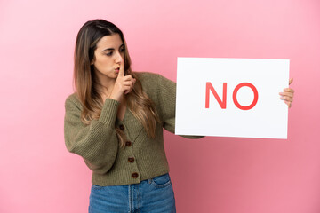Wall Mural - Young caucasian woman isolated on pink background holding a placard with text NO doing silence gesture