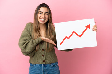 Wall Mural - Young caucasian woman isolated on pink background holding a sign with a growing statistics arrow symbol and  pointing it