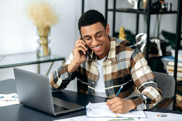 Wall Mural - Smartphone conversation. Successful joyful mixed race hispanic man, stylishly dressed, freelancer or manager, sits at work desk, talking on mobile phone with colleague or client, smiling, takes notes