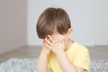 Upset little caucasian boy  in a yellow t-shirt and a large bright room covering his face with his hands and crying