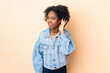 © luismolinero - Young African American woman isolated on beige background listening to something by putting hand on the ear