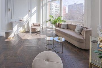 very light and bright interior of luxurious cozy living room with chic soft beige furniture with gold metallic elements, huge window to the floor and wooden parquet