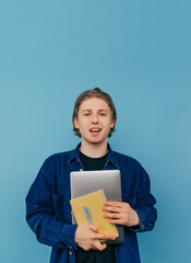 Wall Mural - Vertical portrait of a handsome young man in a shirt with a laptop and a notebook in his hands isolated on a blue background, looking at the camera and smiling.