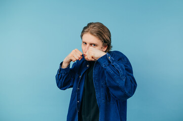 Wall Mural - Young man in a shirt stands in a fighting stance on a blue background and looks at the camera with a serious face. Student boy in boxing rack, isolated.