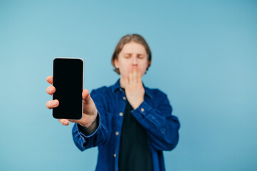 Wall Mural - Shocked guy in a shirt stands on a blue background and shows a smartphone with a black blank screen to the camera, covering his face from surprise. Copy space