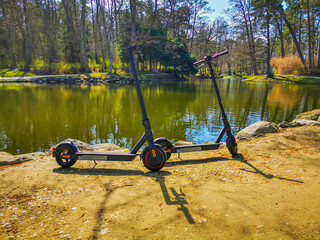 Two electric scooters standing on the footway by the pond in a park