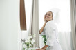 © etonastenka - A lovely young woman in a bathrobe and hair wrapped in a towel looks in the mirror after a shower. Skin care, cleansing and moisturizing concept