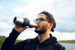 Portrait of young man drinking water from reusable steel thermo bottle on background of blurred cloudy sky. Wearing glasses.