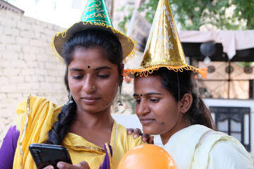 closeup of two young indian female friends wearing party hats looking at the phone outdoors