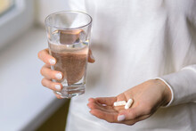 A Woman Holds A Glass Of Water And Two Pills In Her Hands. Young Woman Treats Illness With Pills. Headache Or Stomach Problems And Treatment With Medication. Antibiotic Treatment Concept