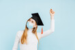 © Shopping King Louie - female graduate in graduation hat and medical mask, student celebrating victory by raising her fists, on blue background