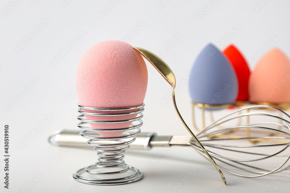 Creative composition with eggs holders, makeup sponges, spoon and whisk on white background, closeup