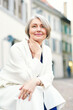 © annanahabed - Outdoor portrait of beautiful and elegant middle age 55 - 60 year old woman, wearing white jacket, sitting on bench outside