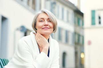 Outdoor portrait of beautiful and elegant middle age 55 - 60 year old woman, wearing white jacket, sitting on bench outside