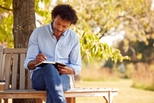 Mature Man Sitting On Park Bench Under Tree Writing In Notebook