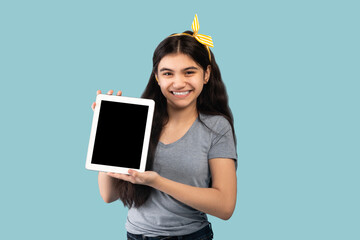 Poster - Cute Indian adolescent girl holding tablet pc with blank screen on blue background, space for app or website design