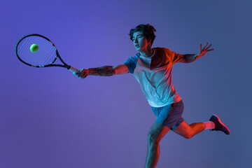 Young caucasian man playing tennis isolated on purple-blue studio background in neon, action and motion concept