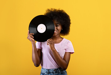 Music lover. Happy african american woman holding vinyl record and covering eye, posing over yellow studio background