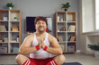 Peaceful thick funny guy in sportswear doing meditation folding hands in pray gesture relaxing in lotus pose sitting on yoga mat at home. Keep calm, dieting, request for strength and endurance