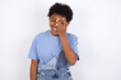 © Roquillo - African American female with afro hair wears jeans overalls over white wall makes face palm and smiles broadly, giggles positively hears funny joke poses