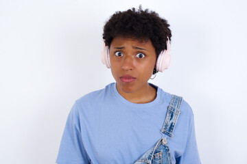 Serious displeased young African American woman with short hair wearing denim overall against white wa looks puzzled at camera being angry wears stereo headphones listens music while walking at street