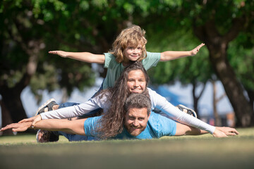 Wall Mural - Cute family portrait. Portrait of a happy smiling family relaxing in park. Family lying on grass in park. Fly concept, little boy is sitting pickaback while imitating the flight.