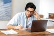 © Kateryna - Concentrated clever young hispanic man wearing glasses, manager, student or freelancer, working at home on laptop, communicates with employees or client via headset while taking notes
