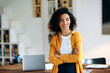 © Kateryna - Portrait of a happy pretty african american girl with curly hair in stylish clothes, freelancer, student or manager, standing near the work desk, with arms crossed, looks to the side, smiling pleasant