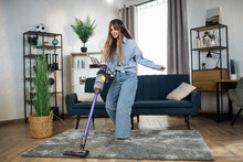 Attractive Woman In Headphones Listening Music And Doing Housekeeping Using Modern Wireless Vacuum Cleaner. Young Housewife Enjoying Cleaning Time With Modern Technology.