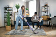 © sofiko14 - Happy caucasian woman listening music in wireless headphones while cleaning carpet with modern vacuum cleaner. Afro american man sitting on sofa and working on laptop.
