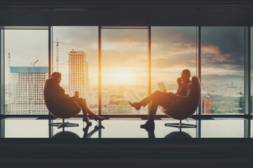 Two business partners sitting in front of each other on elegant armchairs: a businessman with a laptop and a woman entrepreneur with a paper cup of coffee; stunning sunset cityscape outside the window