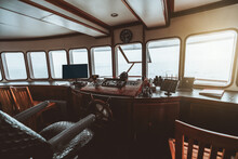 Wide-angle View Of A Captain's Cabin On The Yacht With A Dashboard On The Wooden Basement With Monitors, Navigation Equipment, And Steering Wheel On It; Captain's Armchair In Front Of A Row Of Windows