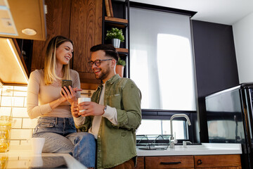 Wall Mural - Beautiful young couple is using a phone and smiling while drinking a coffee in kitchen at home.