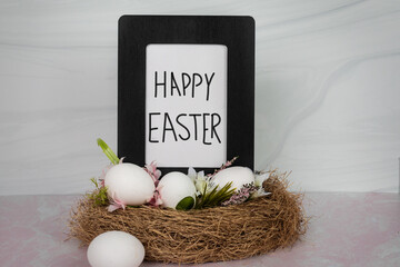 Happy Easter handwritten on white poster in black wood frame sitting in birds nest with white and pink spring flowers and greenery 