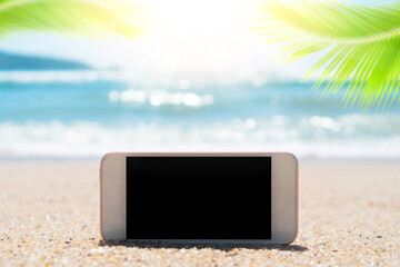 Wall Mural - Smart phone on tropical sand beach with palm leaf and bokeh sunlight wave abstract background. Black copy space of technology and travel nature concept.