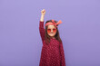 Little fashionable rebel girl dressed in dress, hair band and stylish glasses looks at camera with a grave expression on her face and raised fist up, child defends her rights on purple background