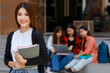 © Bangkok Click Studio - Young and beautiful Asian college student girls holding books, pose to camera with group of friends blur in background against school building. Learning and friendship of teens close friend concept