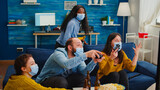 Fototapeta Londyn - People screaming to each other loosing video game playing at new normal party during social pandemic with face mask, keeping distancing against spreading covid 19 virus sitting on couch in living room
