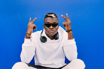 Wall Mural - Young afro American black man wearing a white sweatshirt, sunglasses, comb and headphones on a blue wall looking at camera while doing the victory sign