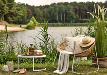 Stylish Composition Of Outdoor Garden On The Lake With Design Rattan Armchair, Coffee Table, Plaid, Pillows, Drinks And Elegant Accessories. Summer Chillout Mood.