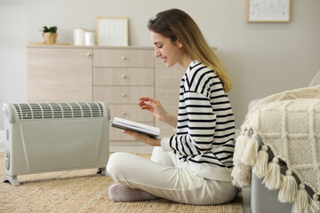 Canvas Print - Young woman with book sitting on floor near electric heater at home